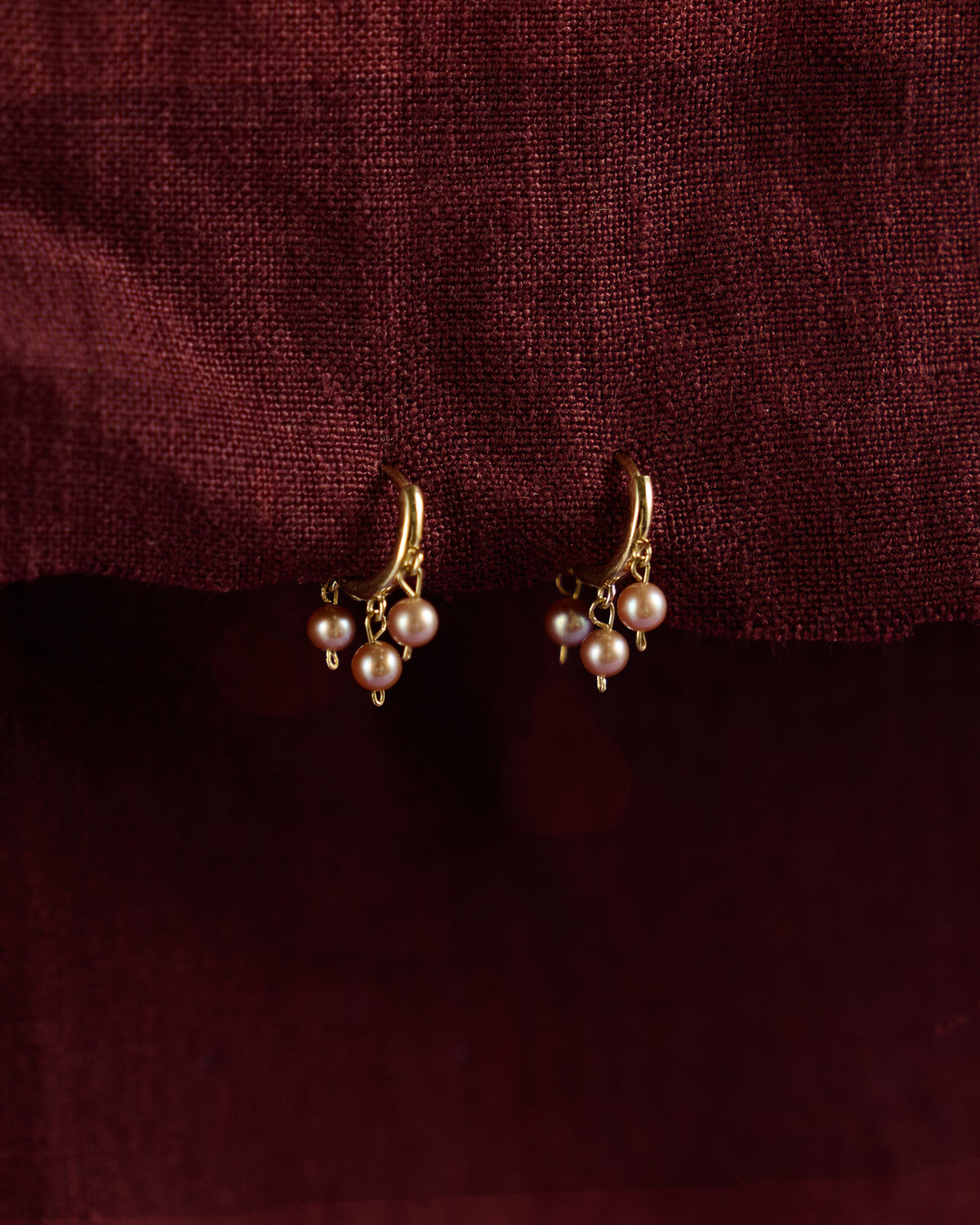 Malar Jhumka Hoops: blush pink pearl jhumka huggie earrings in solid 14k gold on burgundy linen
